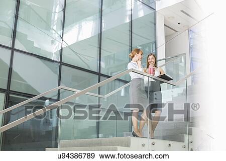 Businesswomen conversing while having coffee on steps in office View Large Photo Image Stock Photo - Businesswomen conversing while having coffee on steps in office. Fotosearch