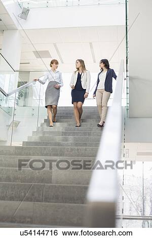 Stock Photograph - Businesswomen conversing while moving down steps in office. Fotosearch