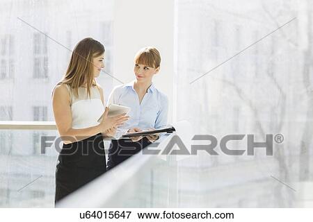 Businesswomen discussing over tablet PC while standing by railing in office View Large Photo Image Stock Photo - Businesswomen discussing over tablet PC while standing by railing in office. Fotosearch