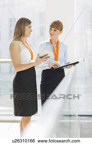 Stock Photograph - Businesswomen discussing over tablet PC while standing by railing in office. Fotosearch