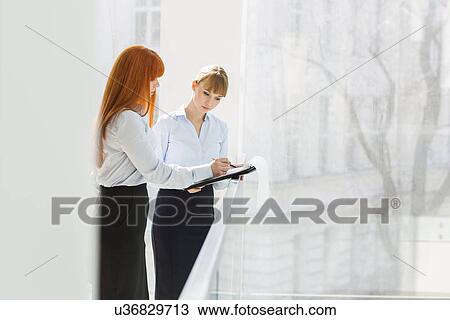 Businesswomen doing paperwork while standing by railing in office View Large Photo Image Stock Image - Businesswomen doing paperwork while standing by railing in office. Fotosearch