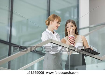 Stock Photography - Businesswomen gossiping while having coffee on steps in office. Fotosearch