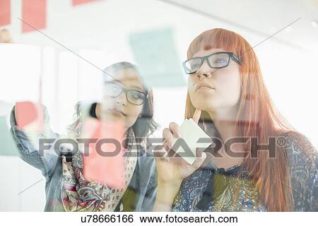 Stock Photograph - Businesswomen reading sticky notes on glass wall in creative office. Fotosearch