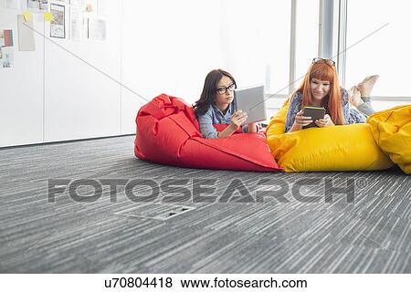Stock Photo - Businesswomen using digital tablets while relaxing on beanbag chairs in creative office. Fotosearch