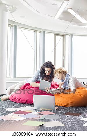 Businesswomen using tablet PC while relaxing on beanbag chairs at creative office View Large Photo Image Picture - Businesswomen using tablet PC while relaxing on beanbag chairs at creative office. Fotosearch