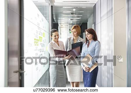 Businesswomen with file folders discussing in office corridor View Large Photo Image Stock Photograph - Businesswomen with file folders discussing in office corridor. Fotosearch