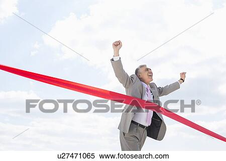 Stock Photography - Cheerful businessman crossing finish line against sky. Fotosearch