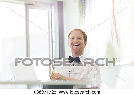 Cheerful businessman sitting at desk in creative office View Large Photo Image Stock Photography - Cheerful businessman sitting at desk in creative office. Fotosearch