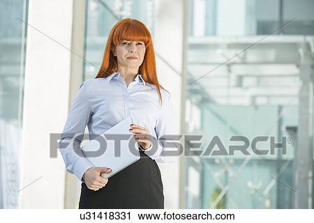 Stock Image - Confident businesswoman looking away while holding laptop in office. Fotosearch