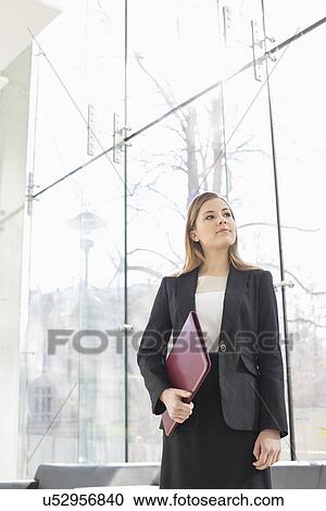 Stock Image - Confident businesswoman looking away while holding folder at office lobby. Fotosearch