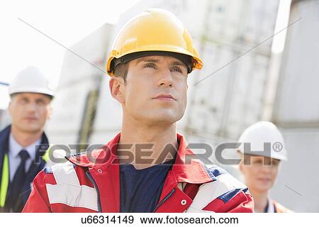 Stock Photo - Confident male worker with colleague in background at shipping yard. Fotosearch