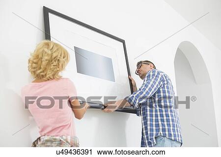 Stock Image - Couple hanging picture frame on wall in new house. Fotosearch