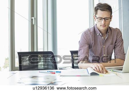 Creative businessman reading file at desk in office View Large Photo Image Stock Photograph - Creative businessman reading file at desk in office. Fotosearch