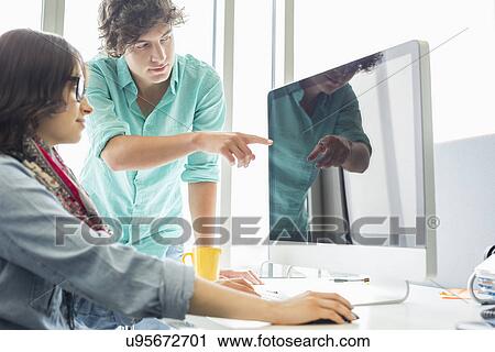 Stock Image - Creative businessman showing something to colleague on desktop computer in office. Fotosearch