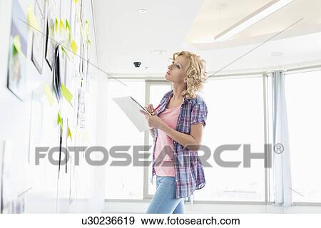 Creative businesswoman looking at papers stuck on wall while writing notes in office View Large Photo Image Stock Photo - Creative businesswoman looking at papers stuck on wall while writing notes in office. Fotosearch
