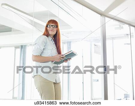 Stock Image - Creative businesswoman looking away while holding files in office. Fotosearch