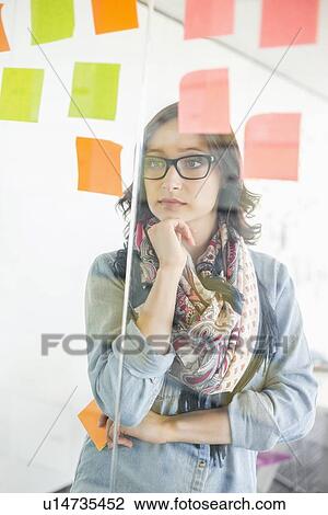 Stock Image - Creative businesswoman reading sticky notes on glass wall in office. Fotosearch
