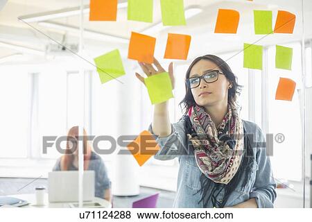 Creative businesswoman reading sticky notes on glass wall with colleague working in background at office View Large Photo Image Stock Photo - Creative businesswoman reading sticky notes on glass wall with colleague working in background at office. Fotosearch