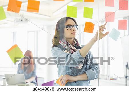 Creative businesswoman reading sticky notes on glass wall with colleague working in background at office View Large Photo Image Stock Photo - Creative businesswoman reading sticky notes on glass wall with colleague working in background at office. Fotosearch