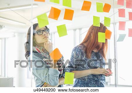 Stock Image - Creative businesswomen reading sticky notes on glass wall in office. Fotosearch