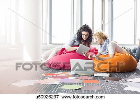 Stock Photo - Creative businesswomen using tablet PC while relaxing on beanbag chairs at office. Fotosearch