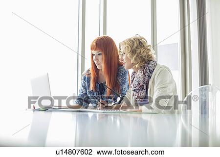 Stock Image - Creative businesswomen working on laptop together in office. Fotosearch