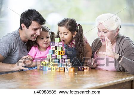 Stock Photography - Cute girl blowing alphabet blocks while family looking at it in house. Fotosearch