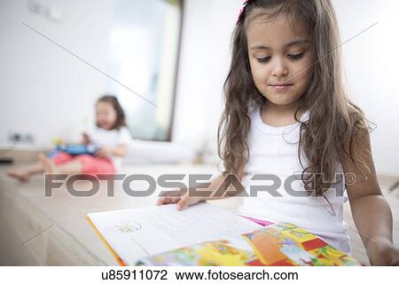 Cute girl reading book with sister in background at home View Large Photo Image Stock Image - Cute girl reading book with sister in background at home. Fotosearch