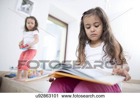 Stock Image - Cute girl reading book with sister playing in background at home. Fotosearch