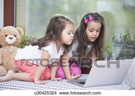 Picture - Cute little sisters using laptop on bed at home. Fotosearch