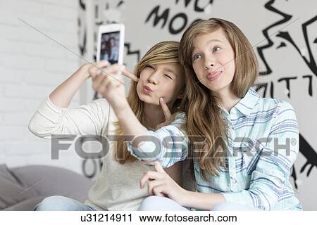 Stock Image - Cute sisters pouting while taking photos with smart phone at home. Fotosearch