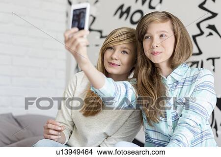 Picture - Cute sisters taking photos with smart phone at home. Fotosearch