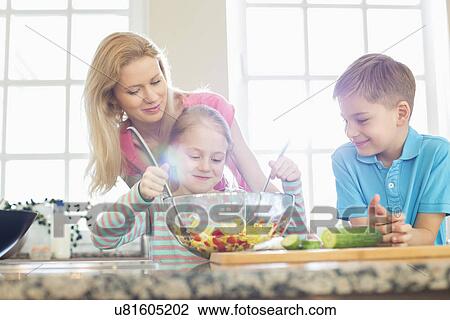 Stock Image - Family looking at girl mixing salad in kitchen. Fotosearch