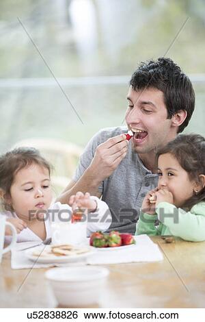 Stock Photograph - Father and daughters eating strawberries with whipped cream at home. Fotosearch
