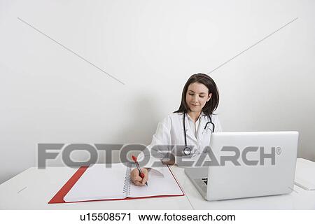 Stock Image - Female doctor writing on binder at desk in clinic. Fotosearch