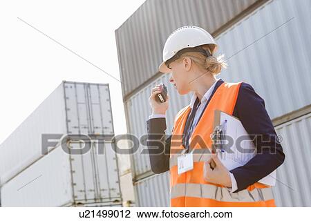Stock Image - Female engineer using walkie-talkie in shipping yard. Fotosearch