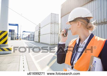 Stock Image - Female engineer using walkie-talkie in shipping yard. Fotosearch