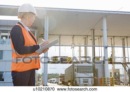 Stock Image - Female worker writing on clipboard while looking at trucks entering in shipping yard. Fotosearch