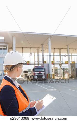 Stock Photo - Female worker writing on clipboard while looking at trucks entering in shipping yard. Fotosearch