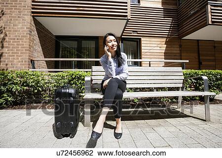 Stock Image - Full length of businesswoman answering cell phone while sitting by luggage on bench against building. Fotosearch
