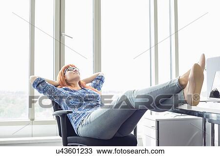 Stock Image - Full-length of businesswoman relaxing with feet up at desk in creative office. Fotosearch