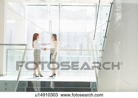 Stock Image - Full-length of businesswomen shaking hands in office. Fotosearch