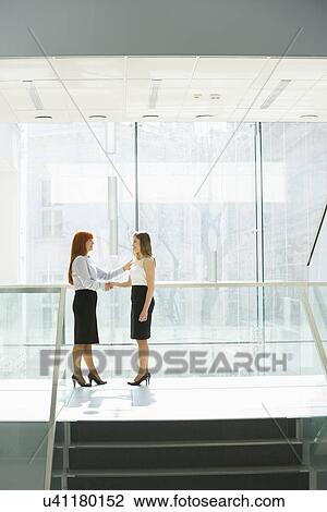 Stock Image - Full-length of businesswomen shaking hands at office hallway. Fotosearch