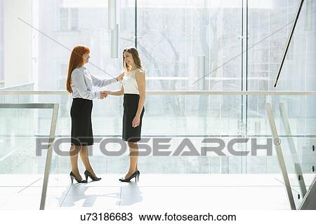 Stock Image - Full-length of businesswomen shaking hands at office hallway. Fotosearch