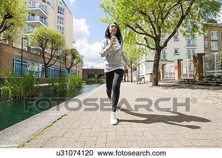 Stock Image - Full length of fit young woman jogging by canal against buildings. Fotosearch
