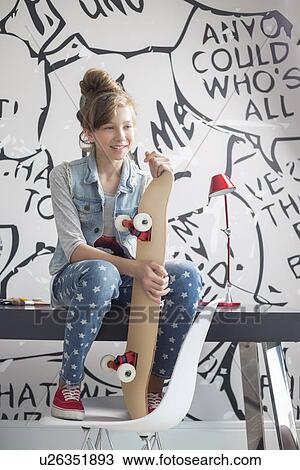 Stock Image - Full-length of girl with skateboard sitting on study table at home. Fotosearch