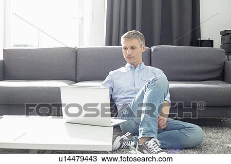 Stock Image - Full-length of man using laptop in living room. Fotosearch
