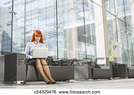 Stock Photograph - Full-length of middle-aged businesswoman using laptop at office lobby. Fotosearch