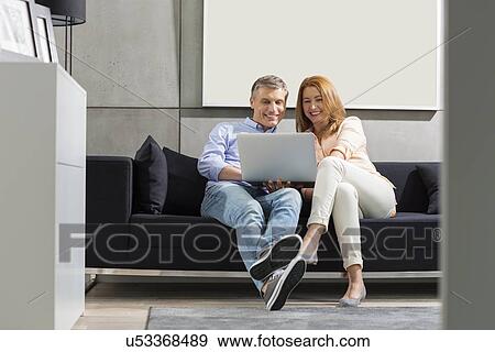 Stock Photo - Full-length of smiling couple using laptop on sofa. Fotosearch