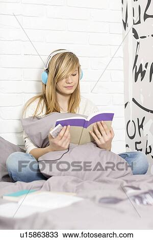 Stock Image - Full-length of teenage girl listening to music while reading book in bedroom. Fotosearch